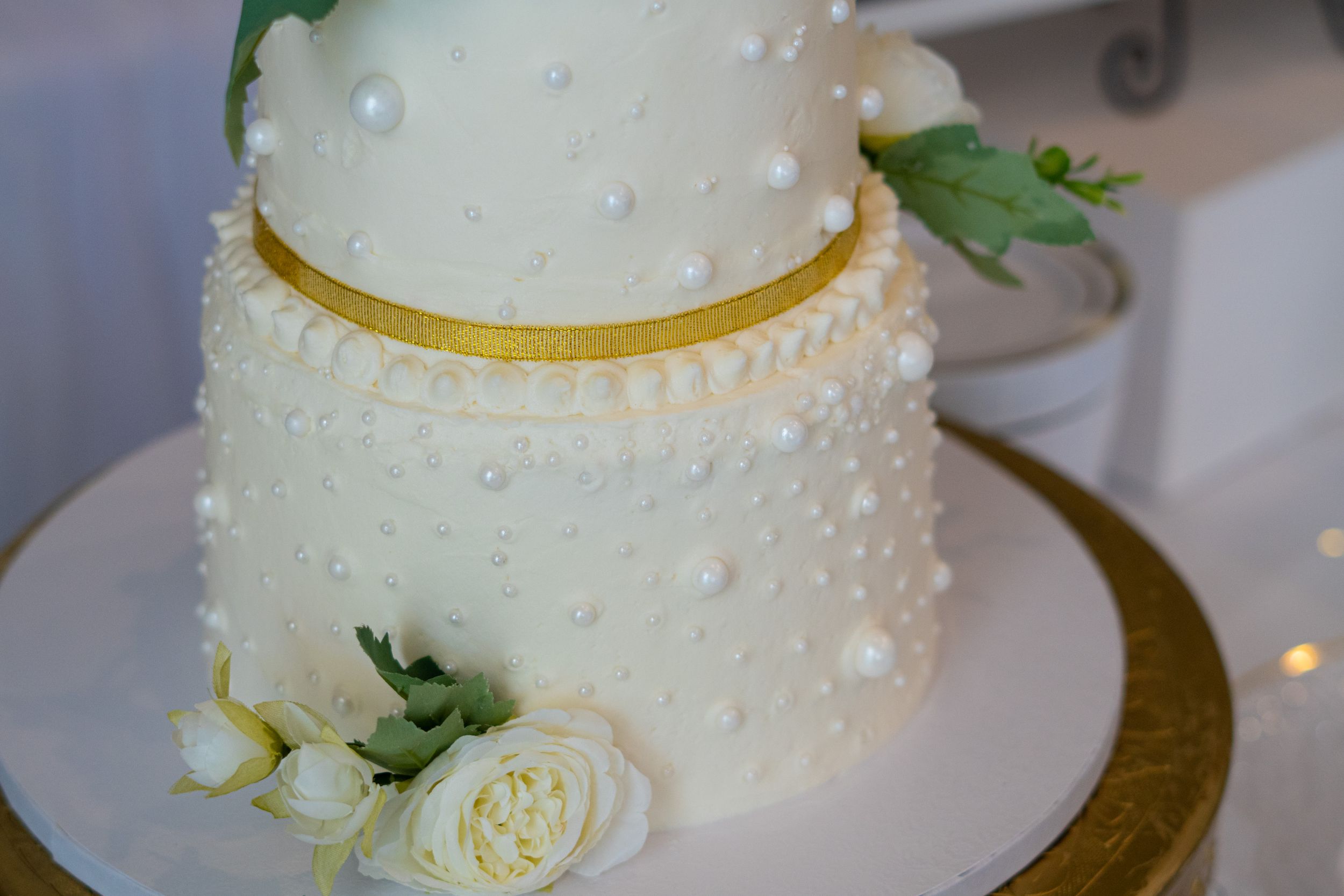 Elegant white wedding cake with pearl details and gold ribbon trim decorated with roses.