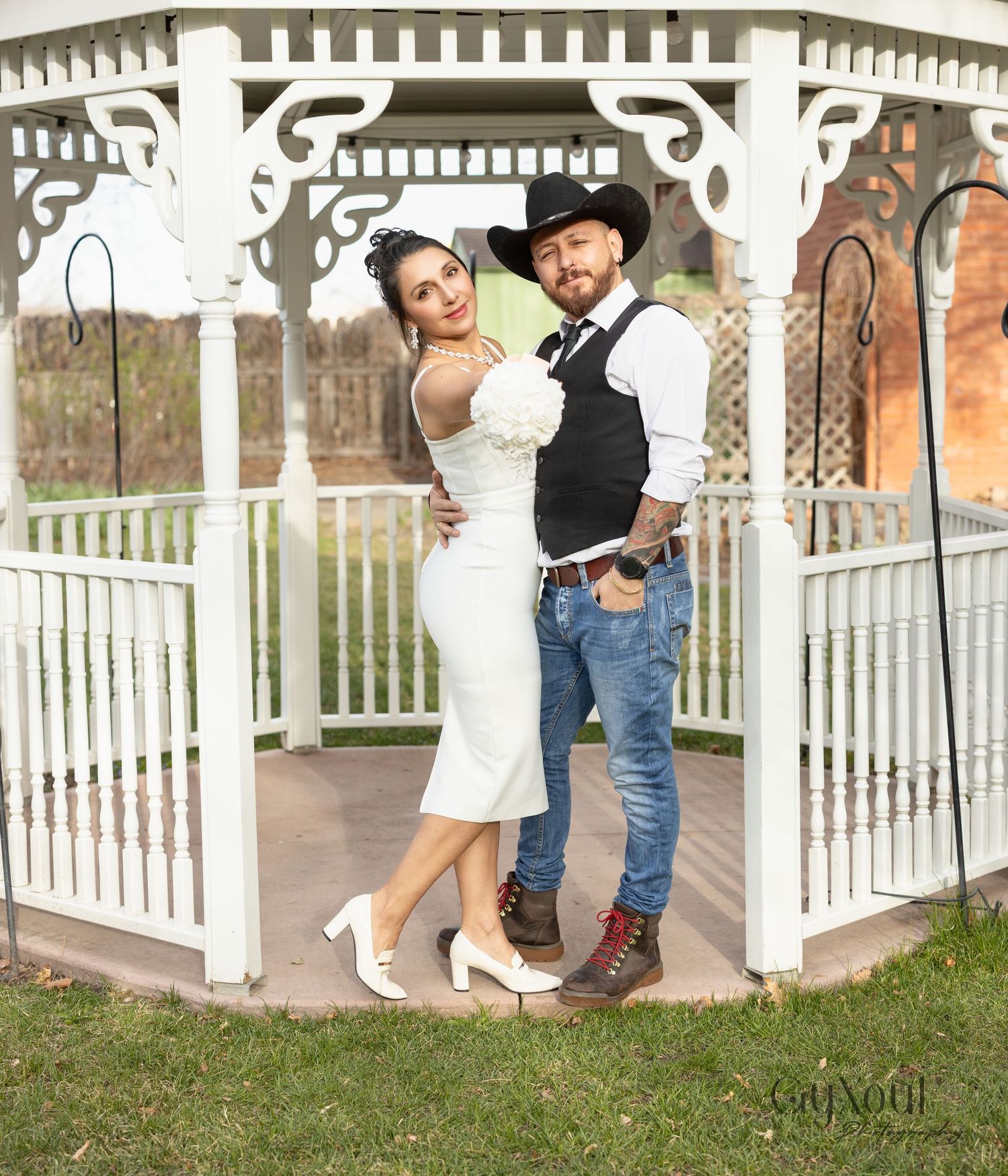 A couple poses in a white gazebo, with the groom in western attire and bride wearing a fitted white dress and heels.