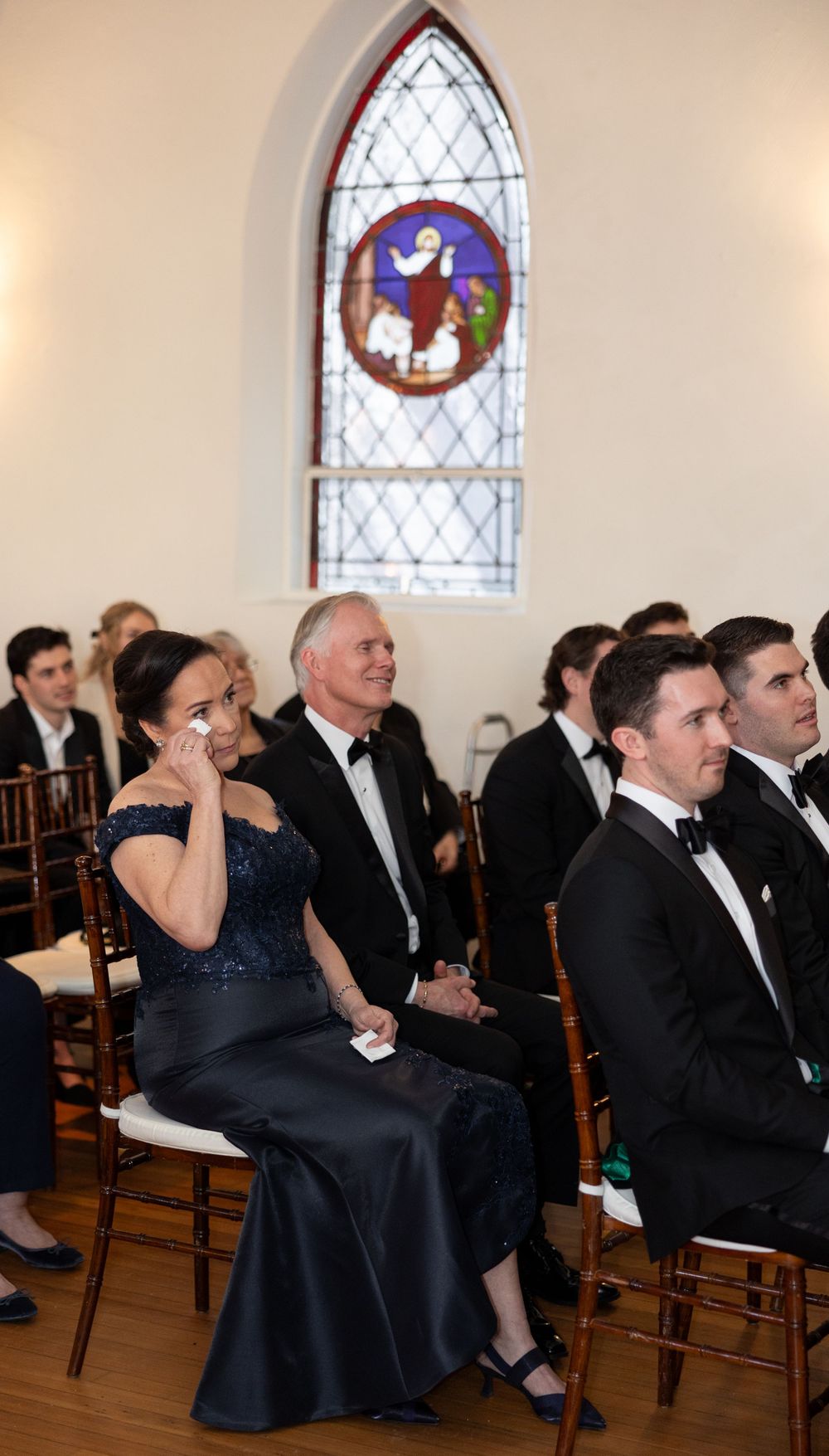 Wedding guests seated in a church with a colorful stained glass window visible in the background.