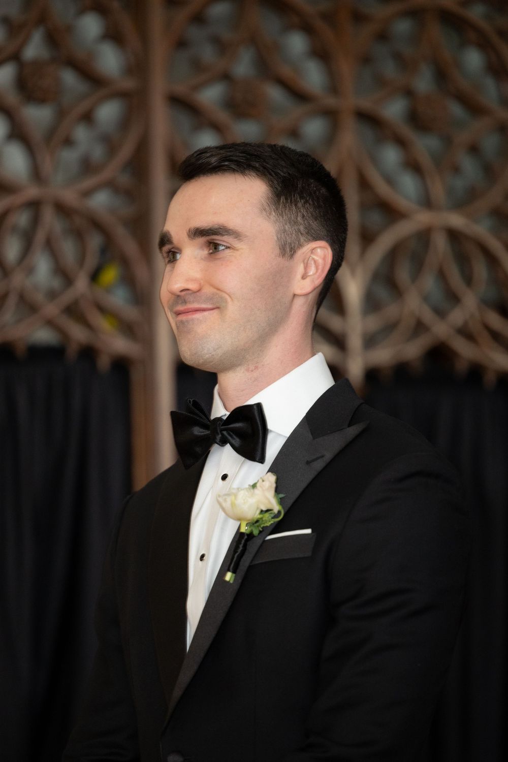 A formal event features a smartly dressed attendee in a black tuxedo and bow tie with boutonniere.