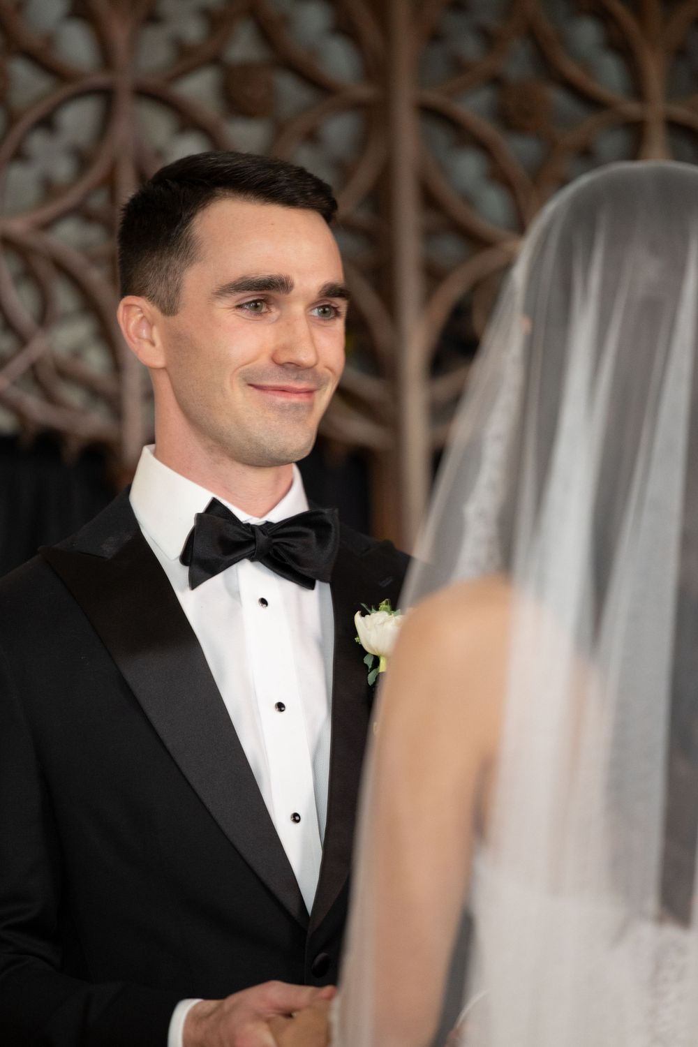 A groom in a black tuxedo and bow tie smiles lovingly during his wedding ceremony.