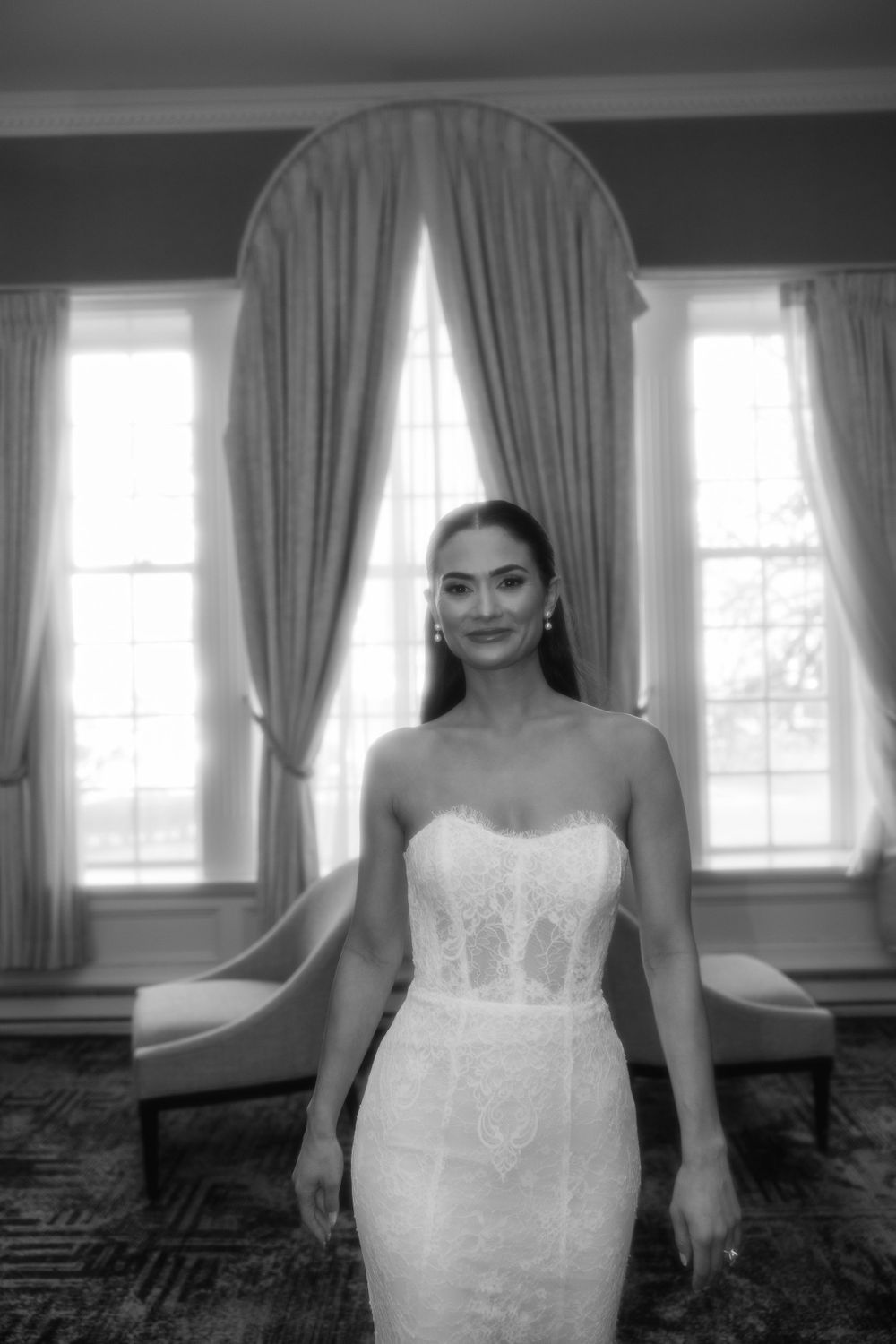 A bride in a fitted white strapless wedding gown stands elegantly in front of tall windows with flowing curtains.