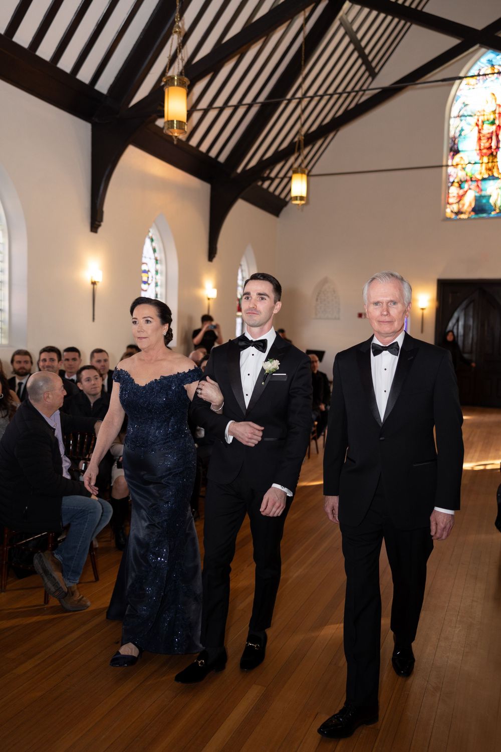 Wedding party members in formal black tie attire walk down the aisle of a historic chapel with vaulted ceilings and stained glass.