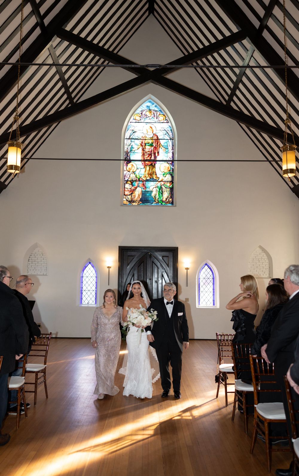 A wedding ceremony in a chapel with stunning stained glass windows and wooden beams casting dramatic lighting.