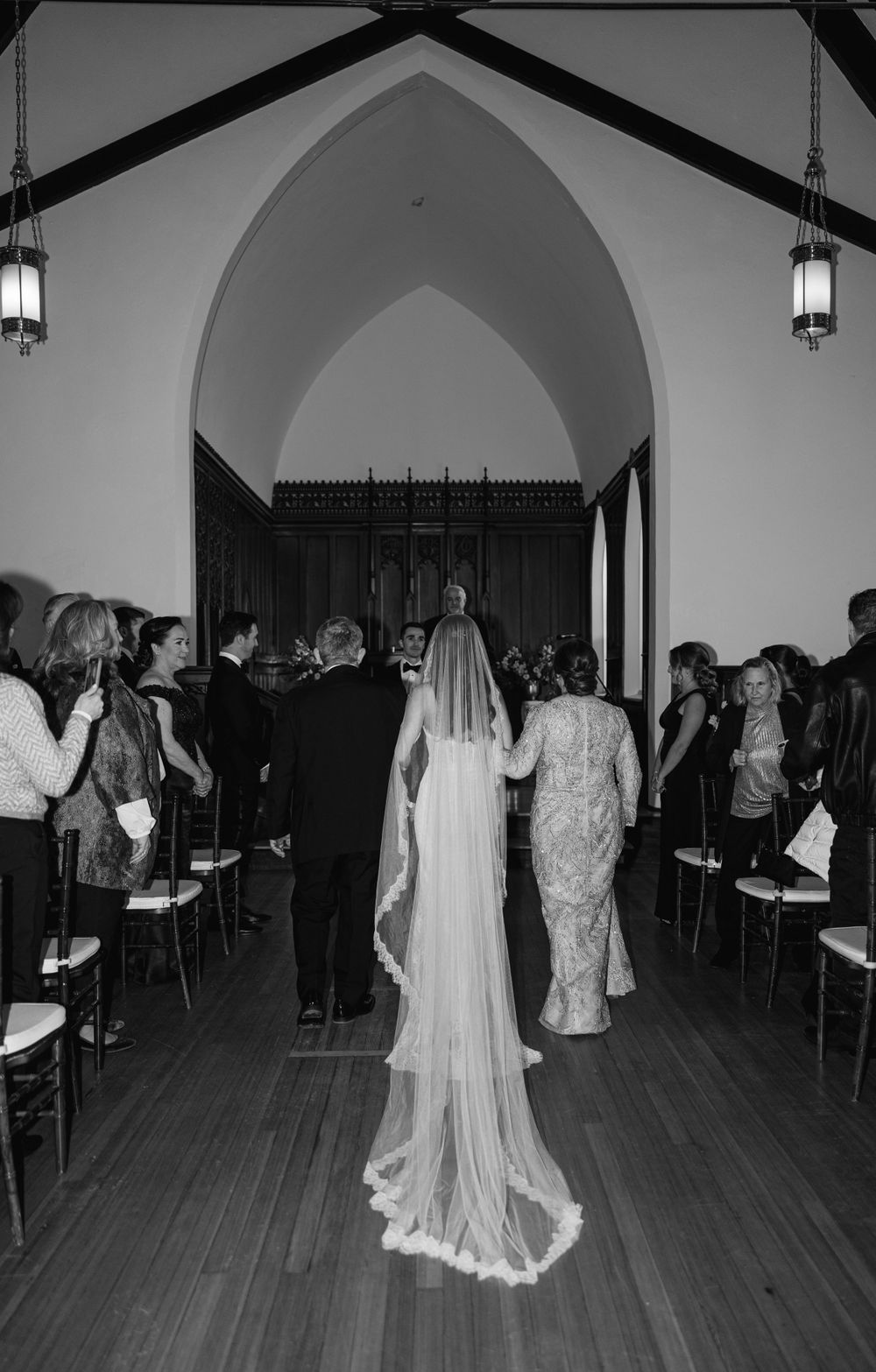 Bride in flowing veil walks down church aisle with guests seated on either side.