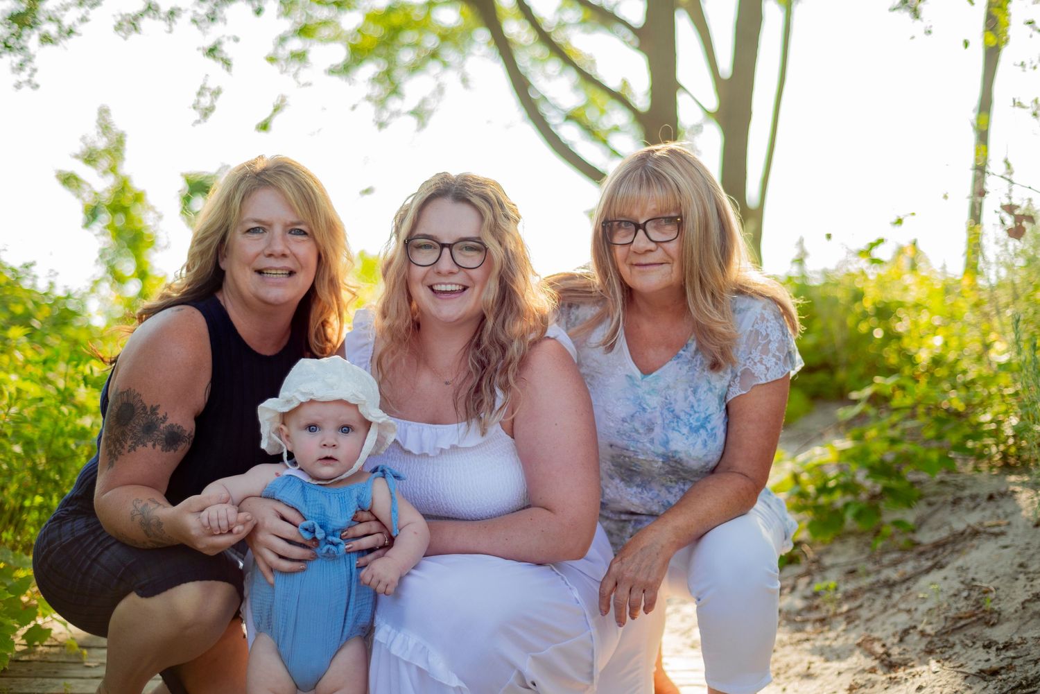 Four generations gather outdoors in a natural setting with sunlight and greenery in the background.