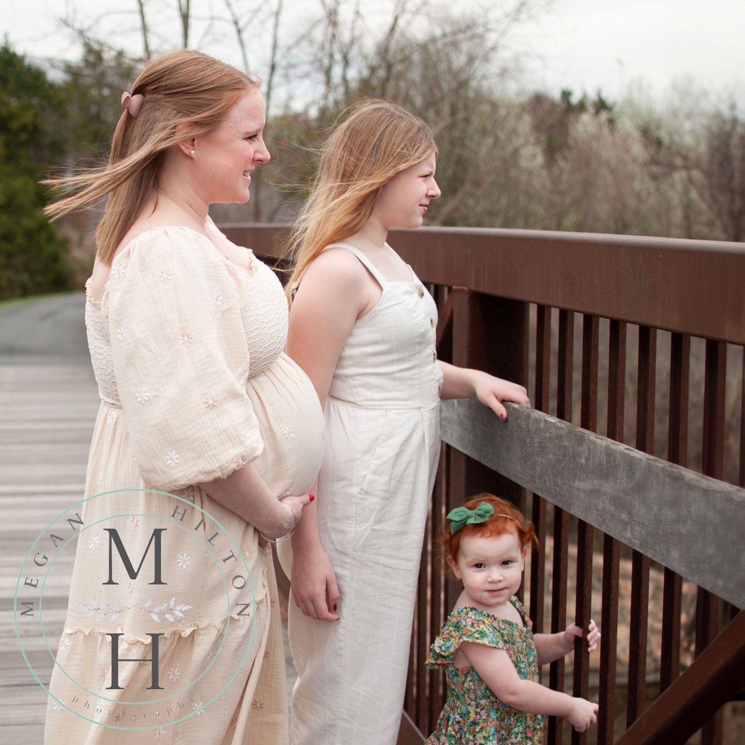 A pregnant mom and her two children stand at a wooden railing on a bridge while a small child peeks through the slats.