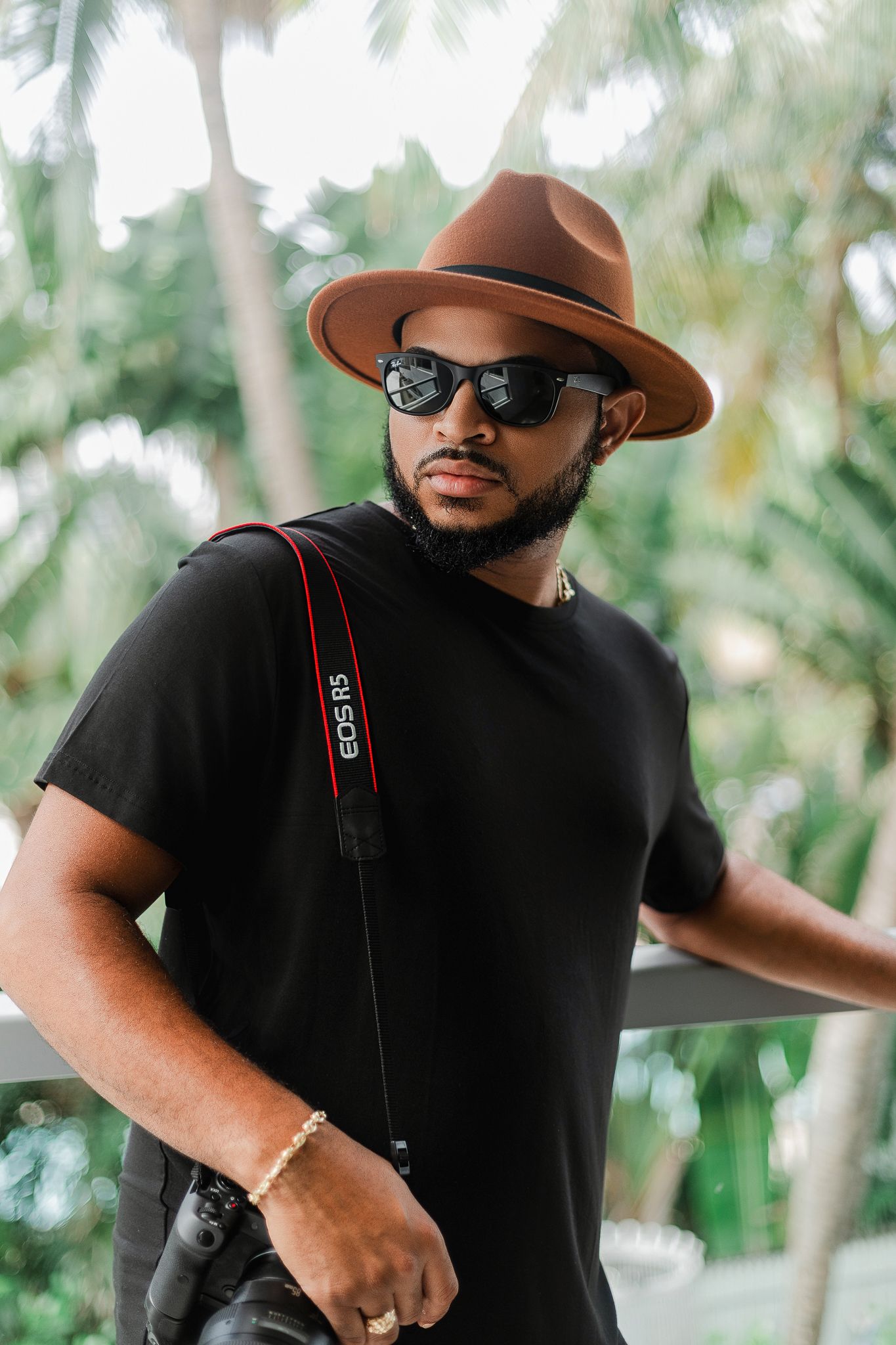A stylish person in a brown fedora and black t-shirt poses against a tropical background.