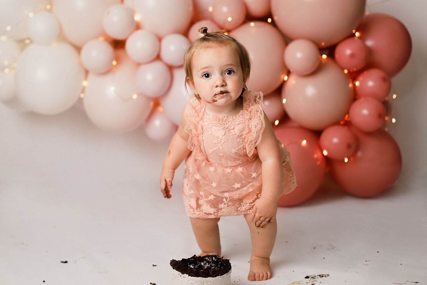 A baby in pink lace outfit sits on floor with pink balloon arch backdrop during cake smash photo session.