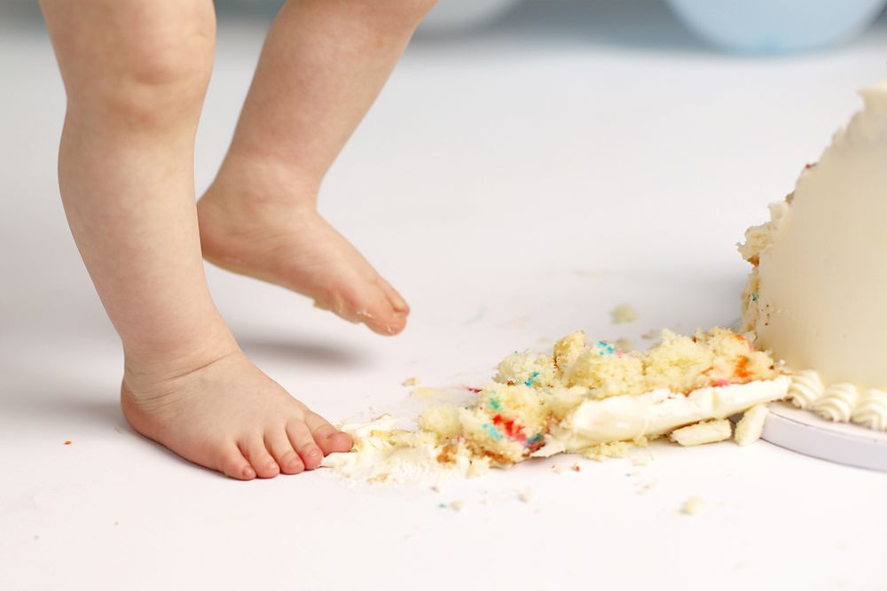 Small bare baby feet standing near scattered cake crumbs and frosting on a light-colored floor.