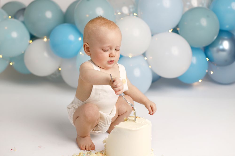 Baby in white outfit sits near birthday cake with blue and white balloon arch backdrop for first birthday celebration.