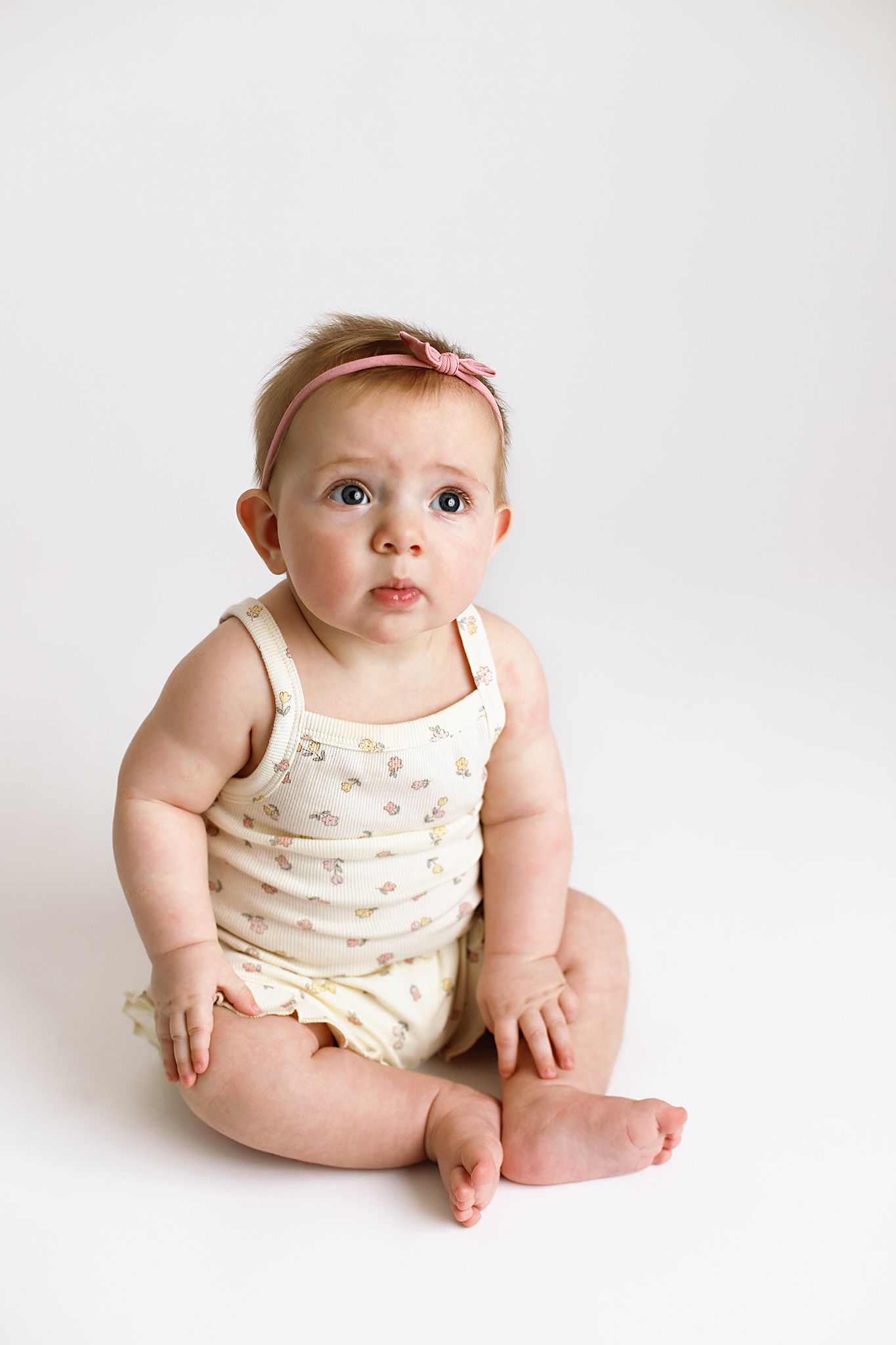 Baby in floral tank top outfit sitting on white background with a pink hair bow.