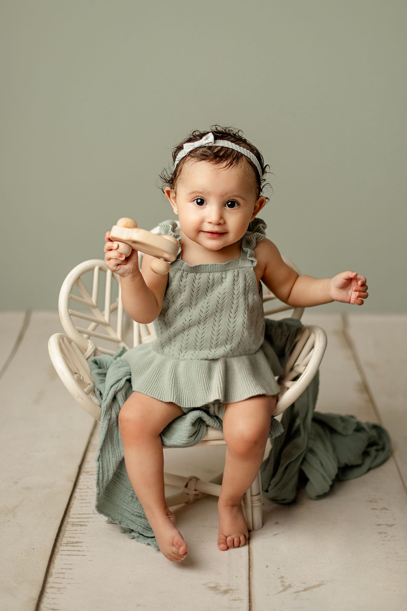 Cute baby girl in sage green knit romper smiling while sitting in a small white rattan chair holding a wooden toy.
