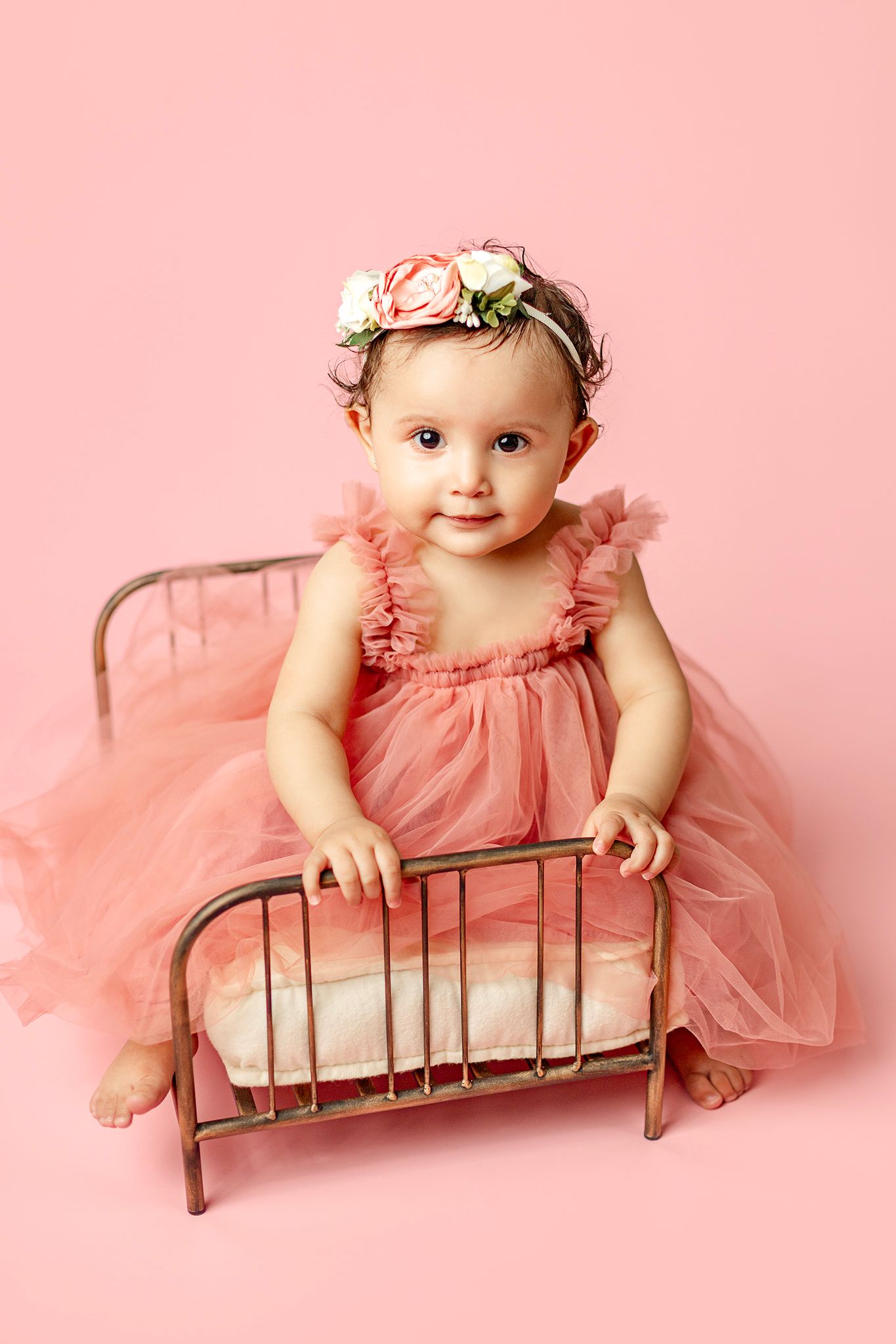 Adorable baby girl in pink tulle dress and floral crown sitting in a miniature metal bed on pink background.