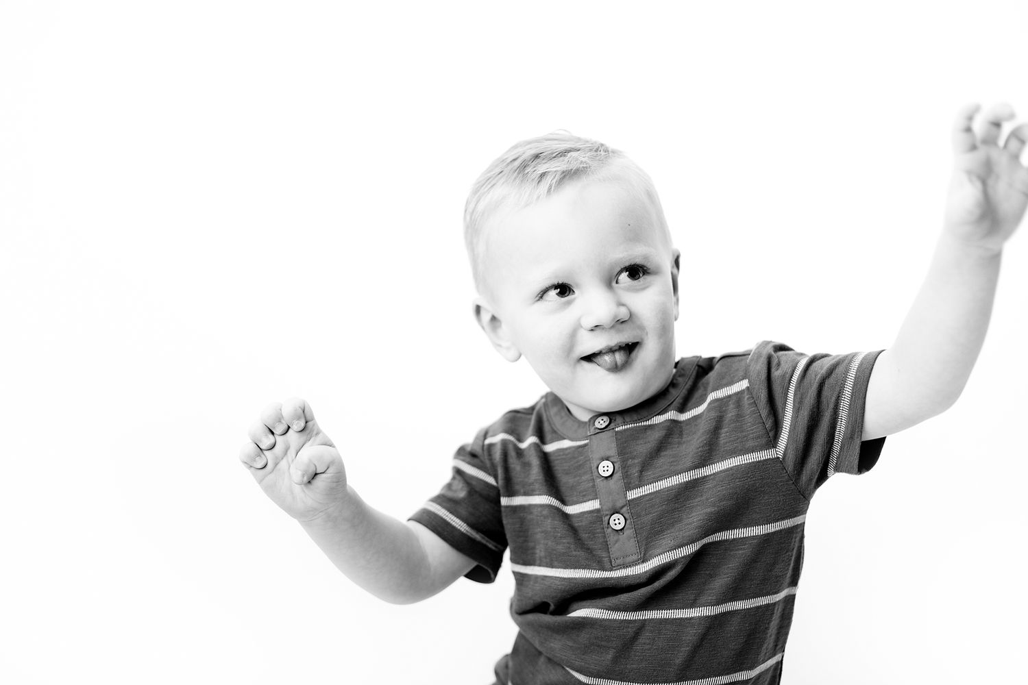 Black and white photo of a happy toddler boy in striped shirt with arms raised joyfully.