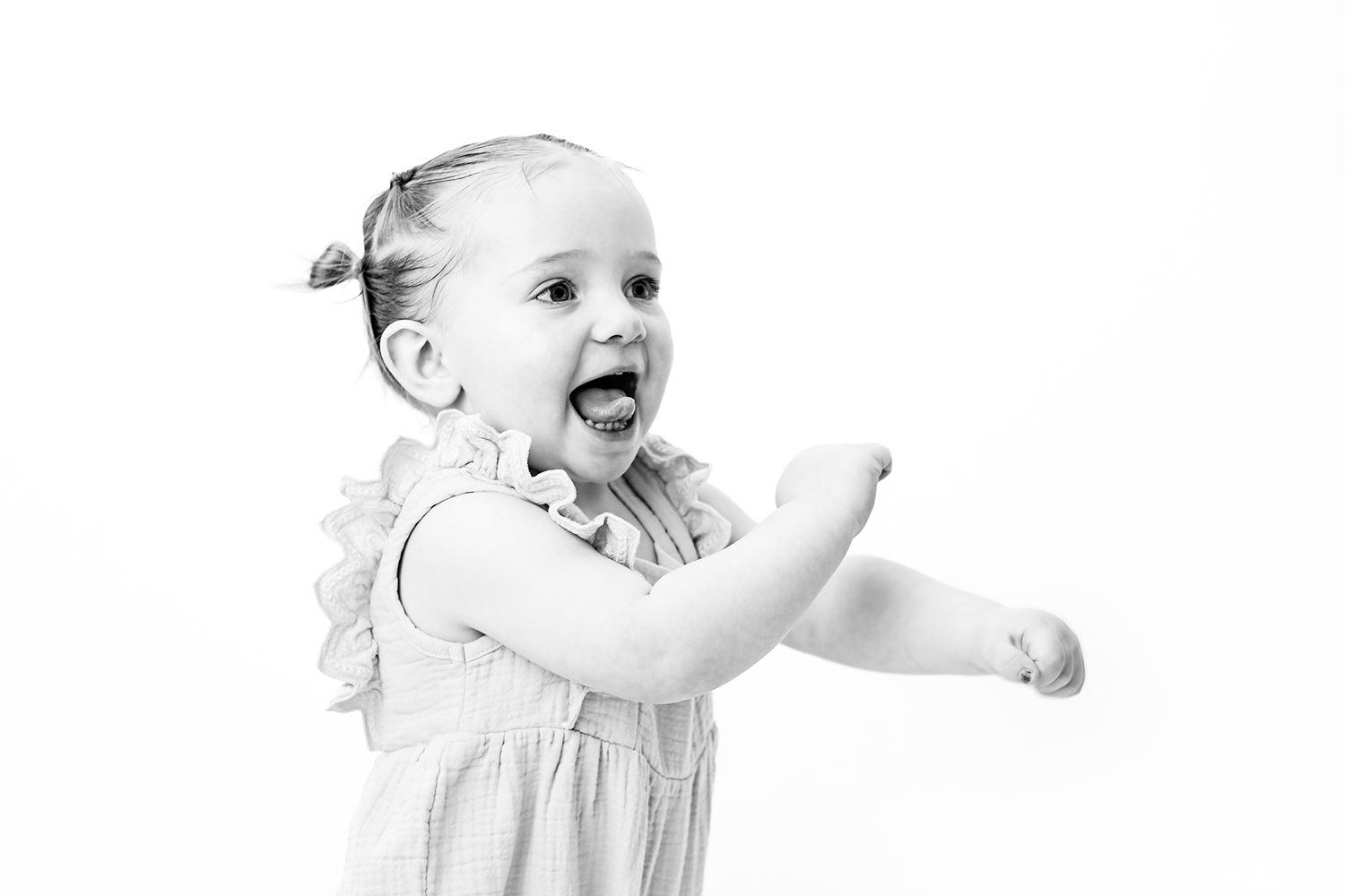 Joyful toddler girl in white dress laughing and reaching out arms in black and white portrait photo.