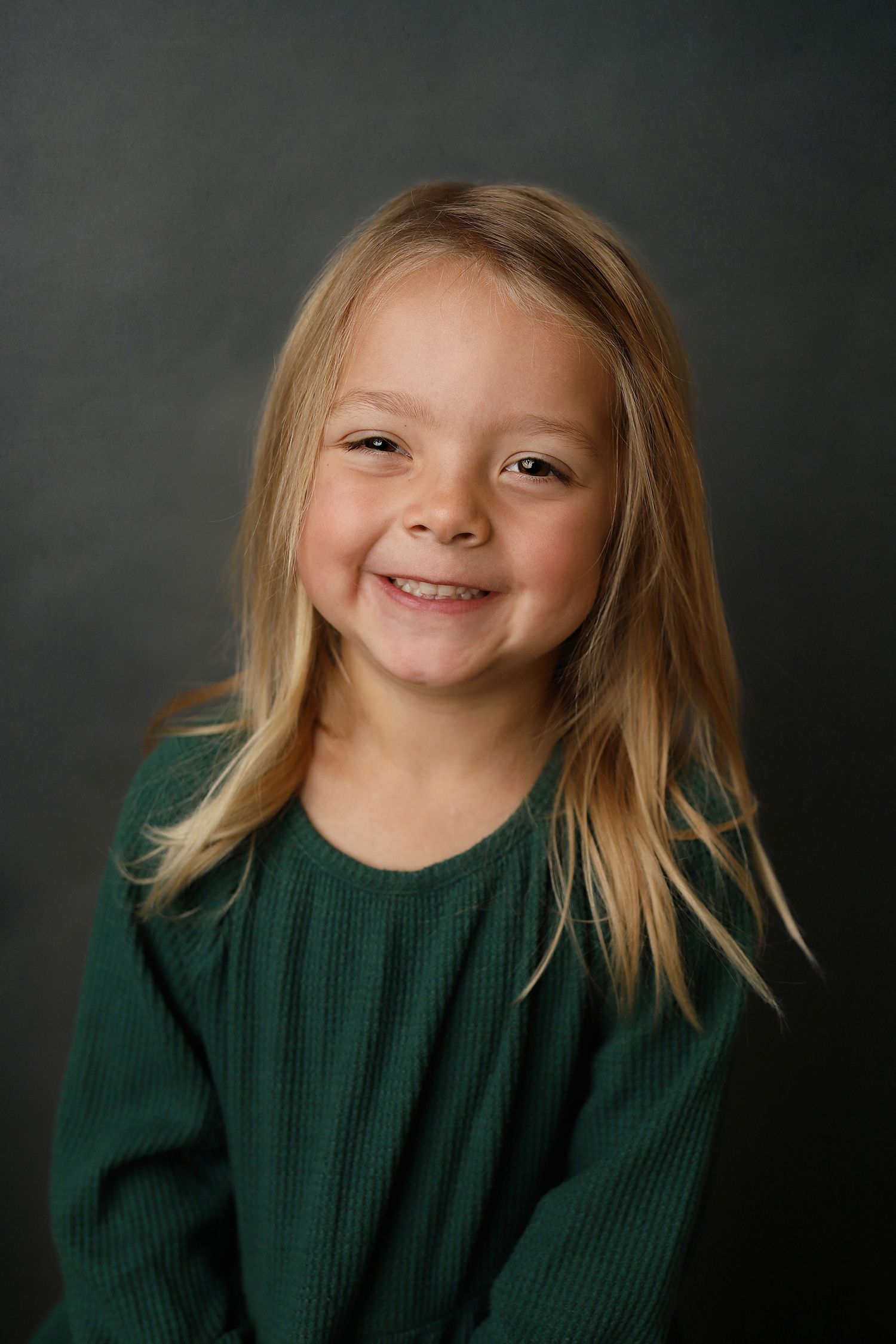 Portrait of a child wearing a dark green sweater smiling warmly against a dark gray background.