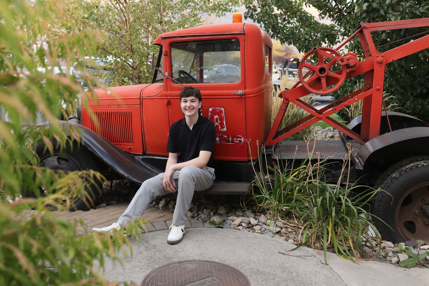 Someone sits casually on the vintage red truck surrounded by greenery in an outdoor setting.