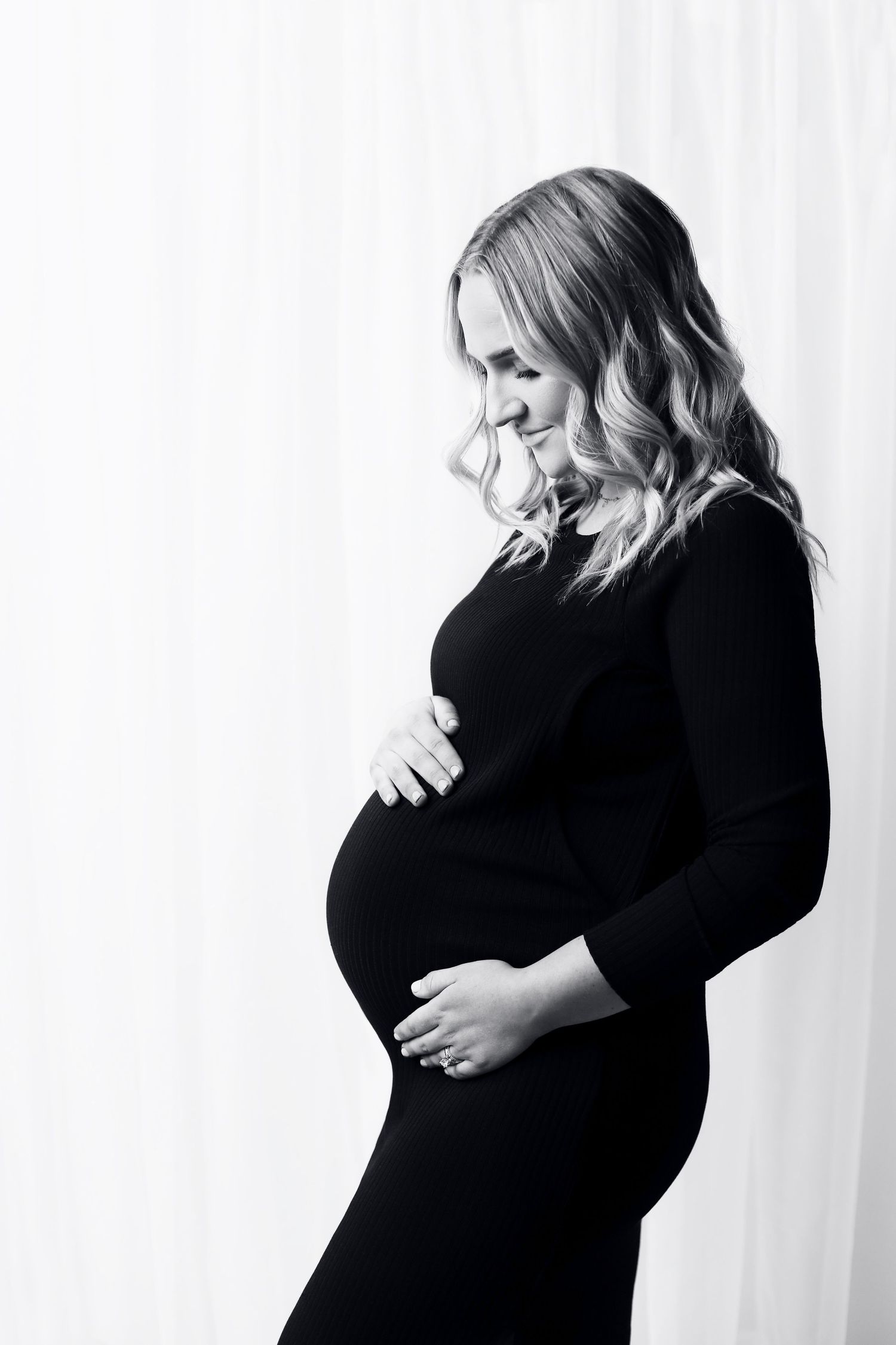 Black and white maternity photo of expectant mother in black dress posed in profile against white background.