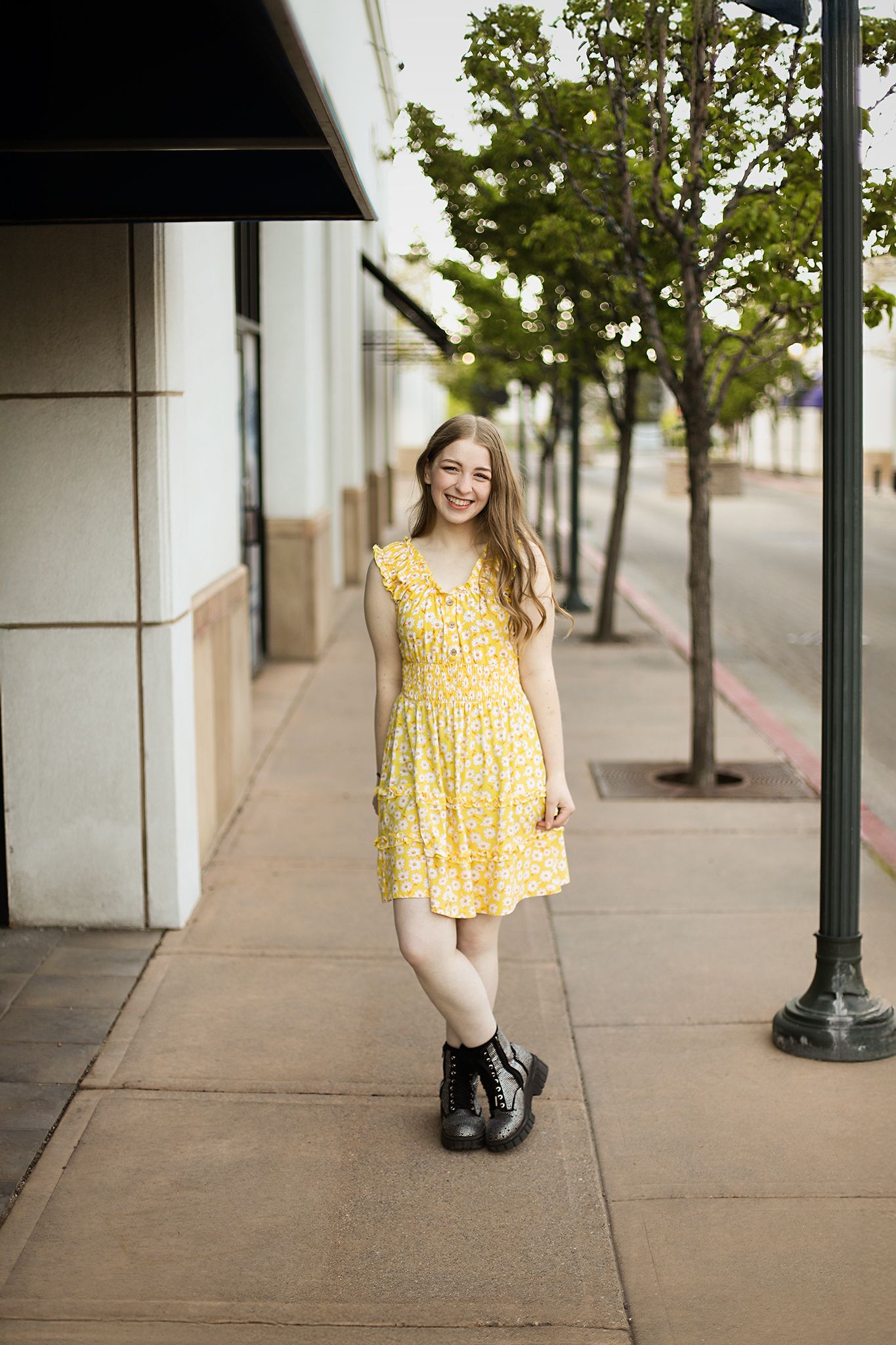 A person wearing a yellow floral sundress and black boots stands on a tree-lined sidewalk outside a building.