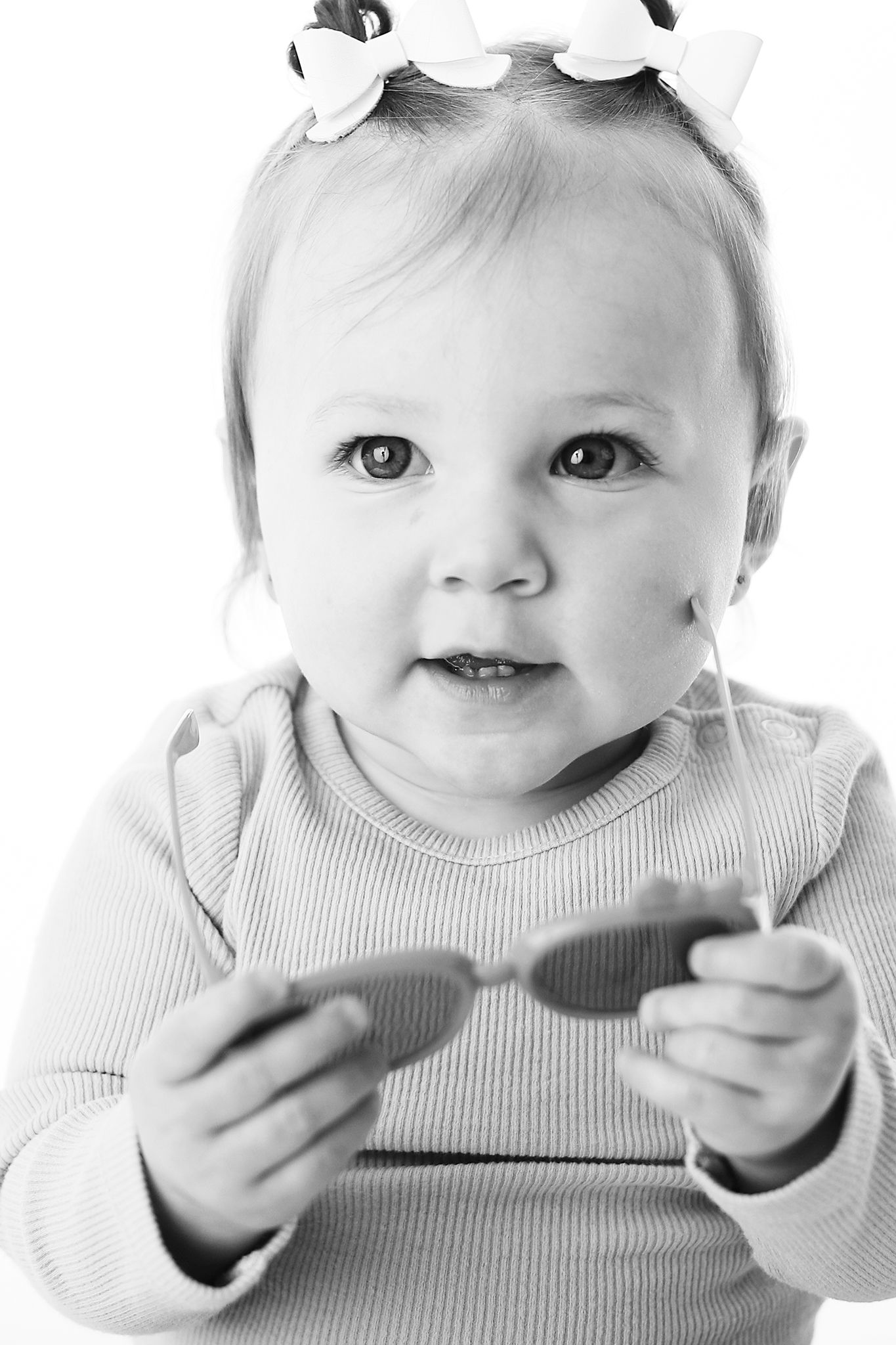 Black and white portrait of a little one with a white bow wearing a ribbed sweater while holding something in both hands.