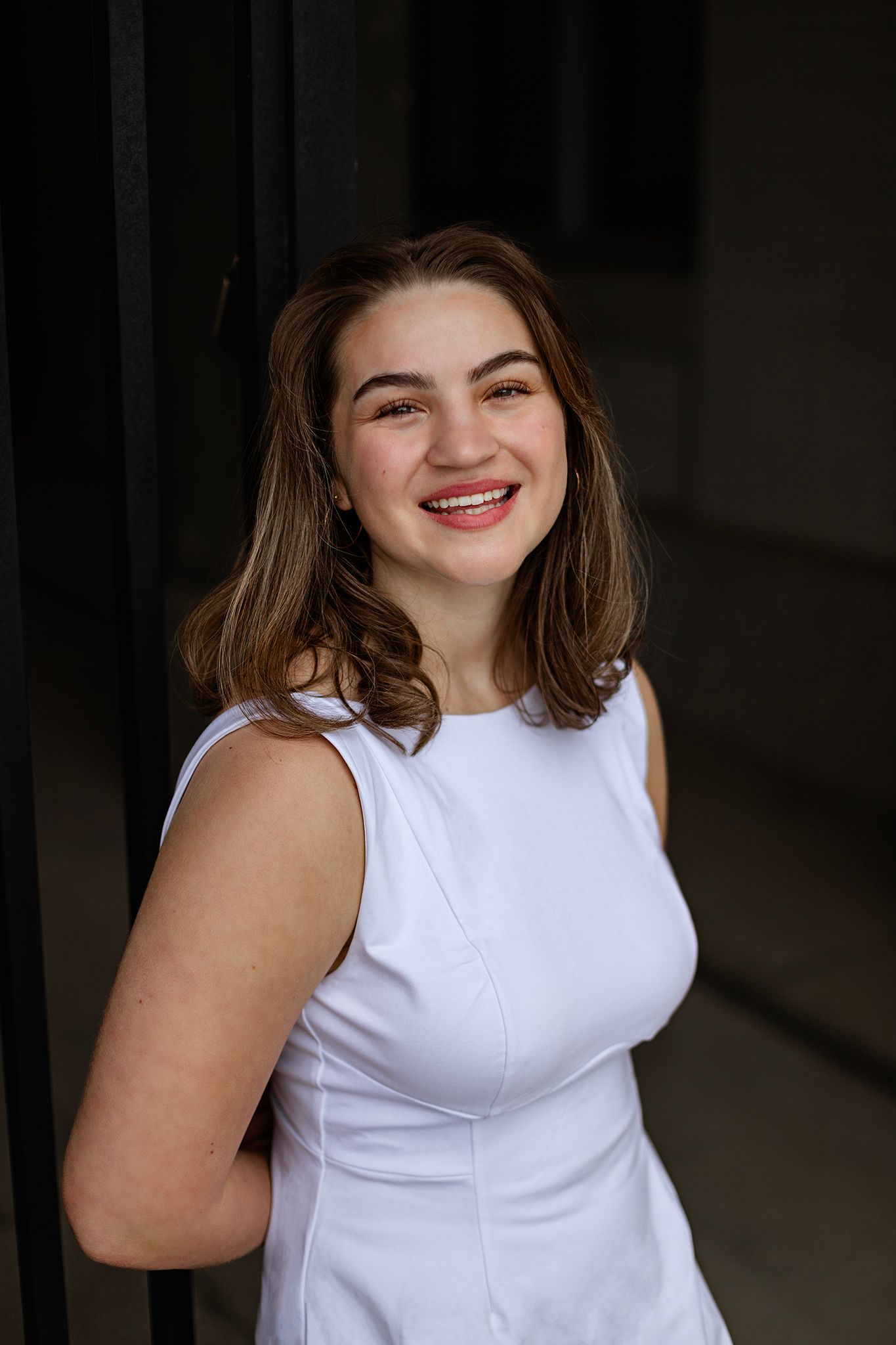 Young woman in white sleeveless dress smiling confidently against dark background.