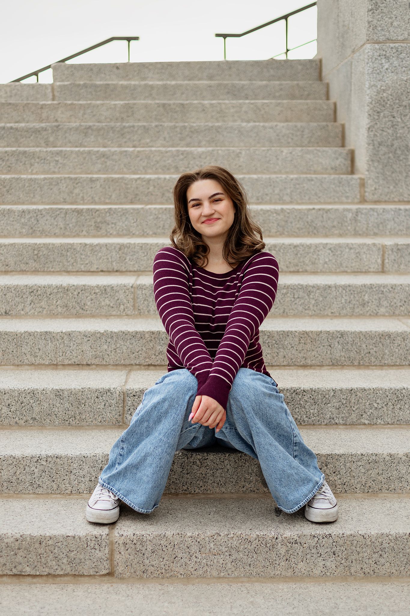 Young woman in striped top and wide-leg jeans sitting on granite steps, smiling confidently at camera.