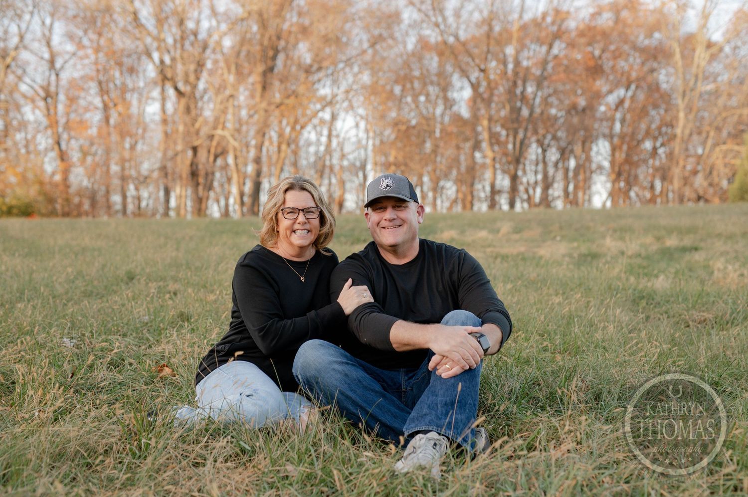 Couple sitting close together on grassy hill during autumn outdoor photoshoot.