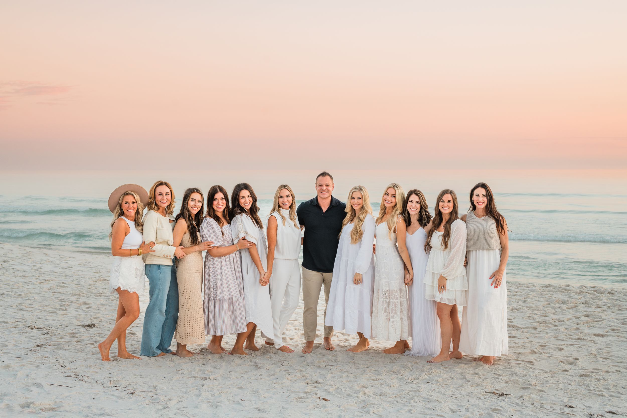 A group in coordinated white and neutral outfits poses together on a beach at sunset with ocean waves in the background.