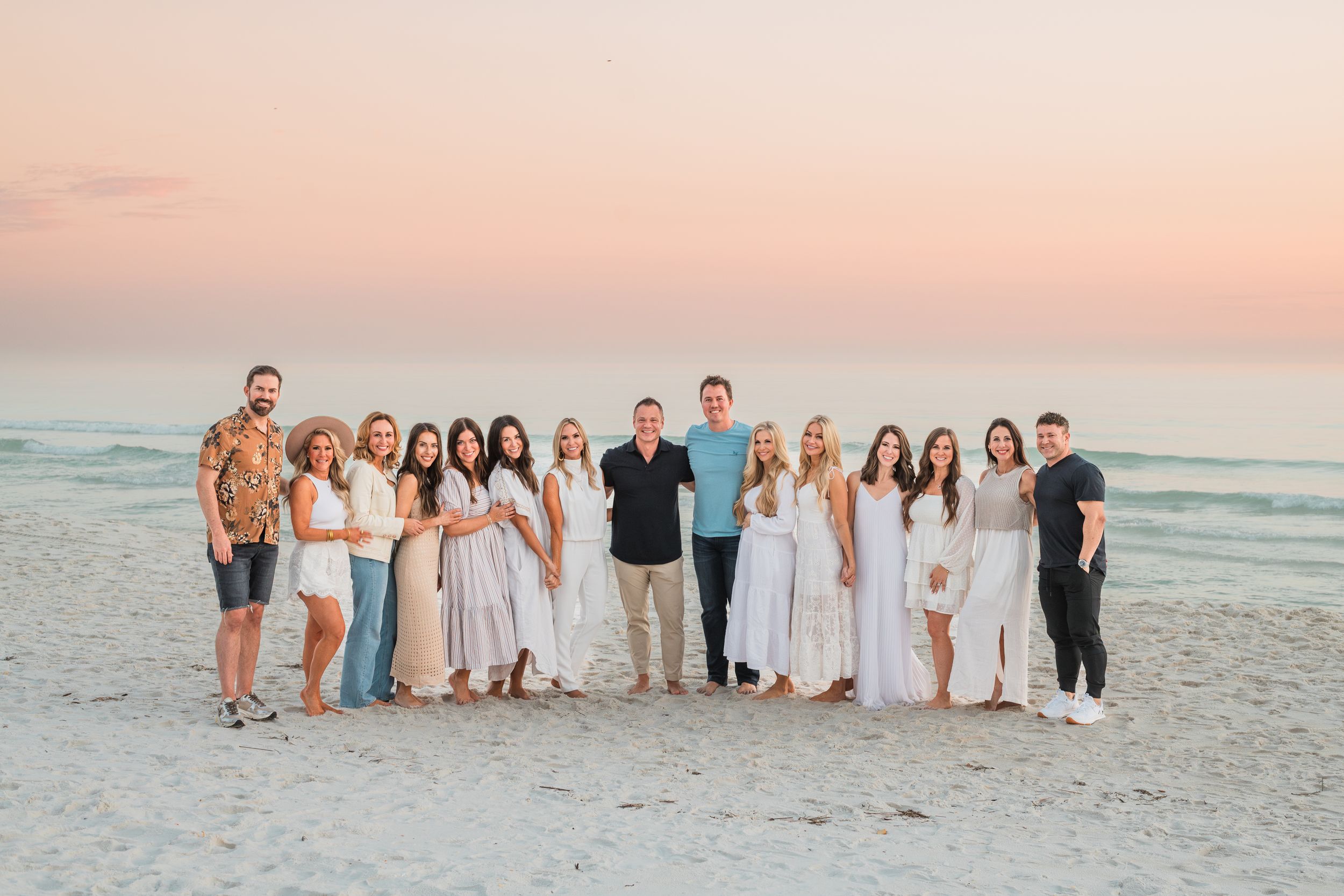 A group dressed in white and neutral colors poses together on a beach at sunset with ocean waves in the background.