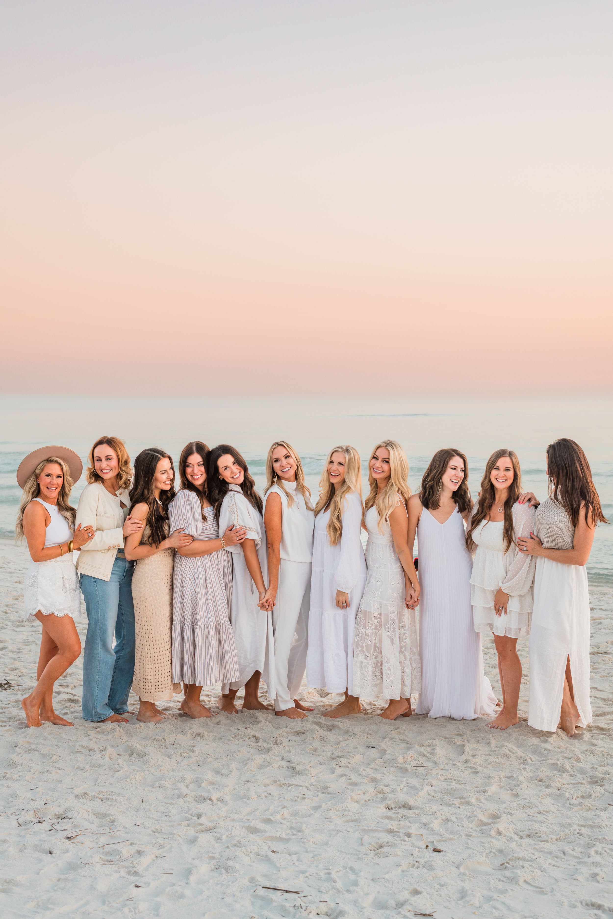 Group in coordinated white and neutral outfits posing together on a beach at sunset with a pastel pink sky.