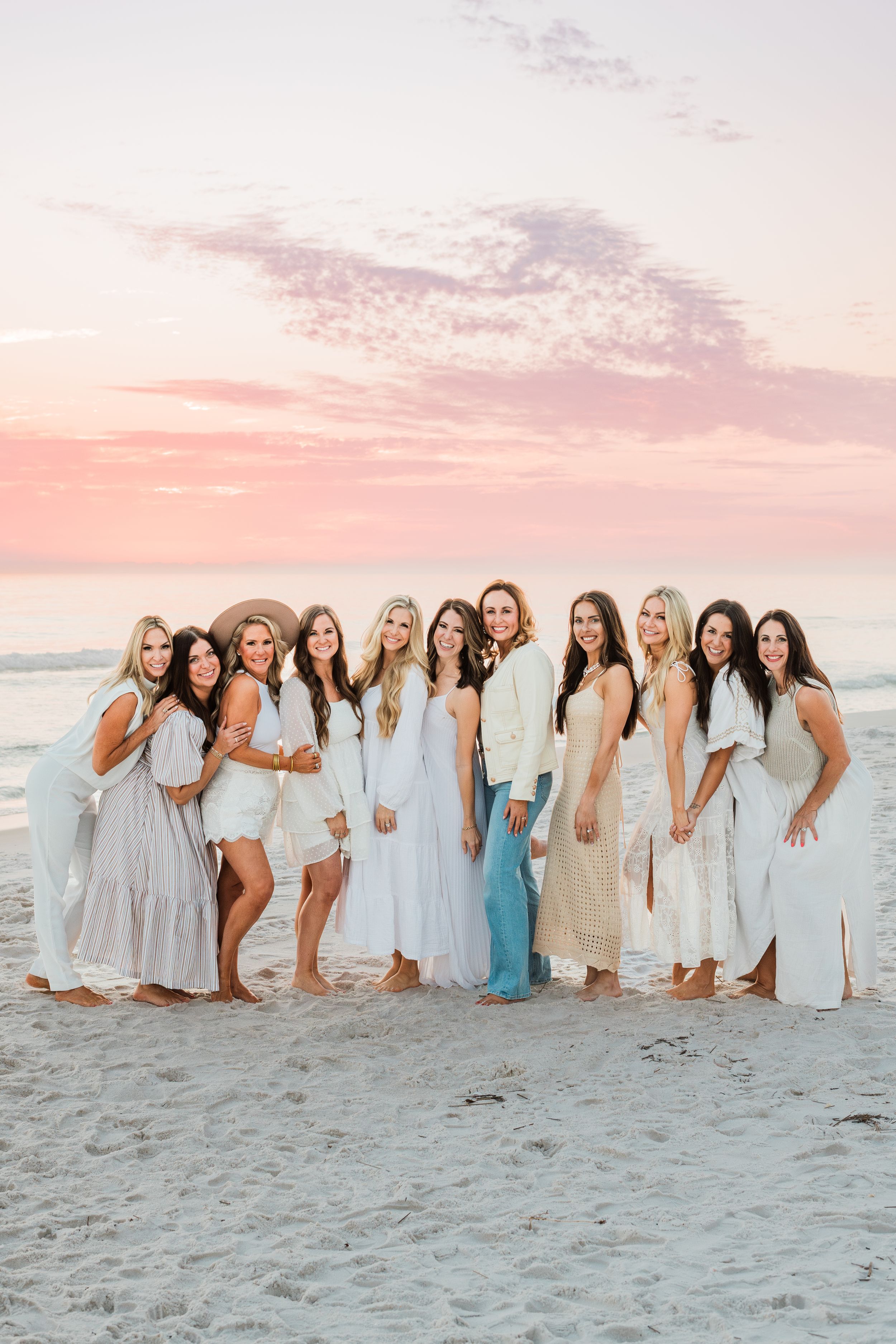 A group of friends in coordinating beige and white dresses pose together on a beach at sunset with pink skies.
