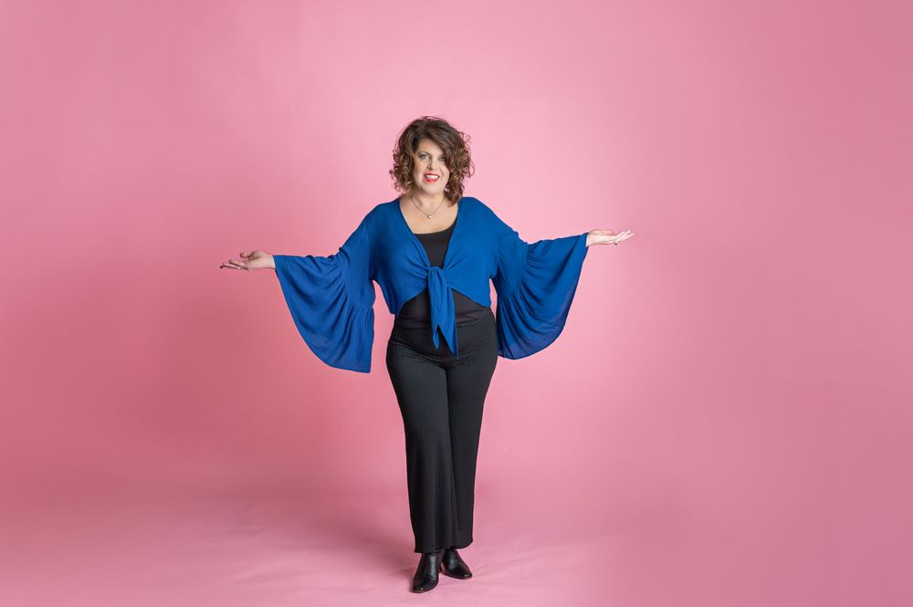 Figure in dramatic royal blue bell-sleeved top with tie front and black pants poses with arms outstretched against pink backdrop.