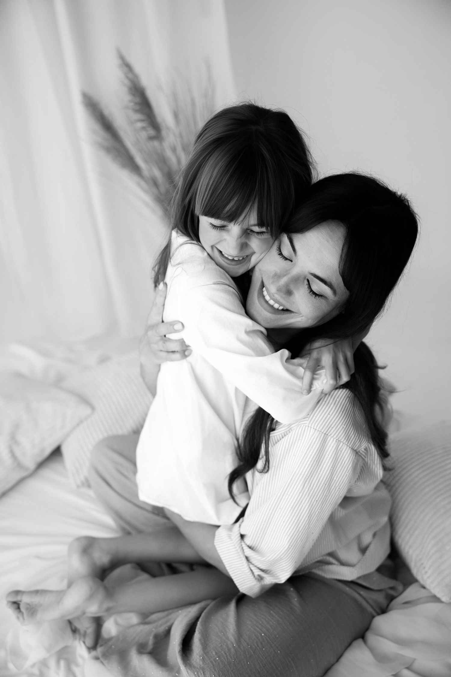 A black and white photo of a smiling mother hugging her laughing daughter on a bed.