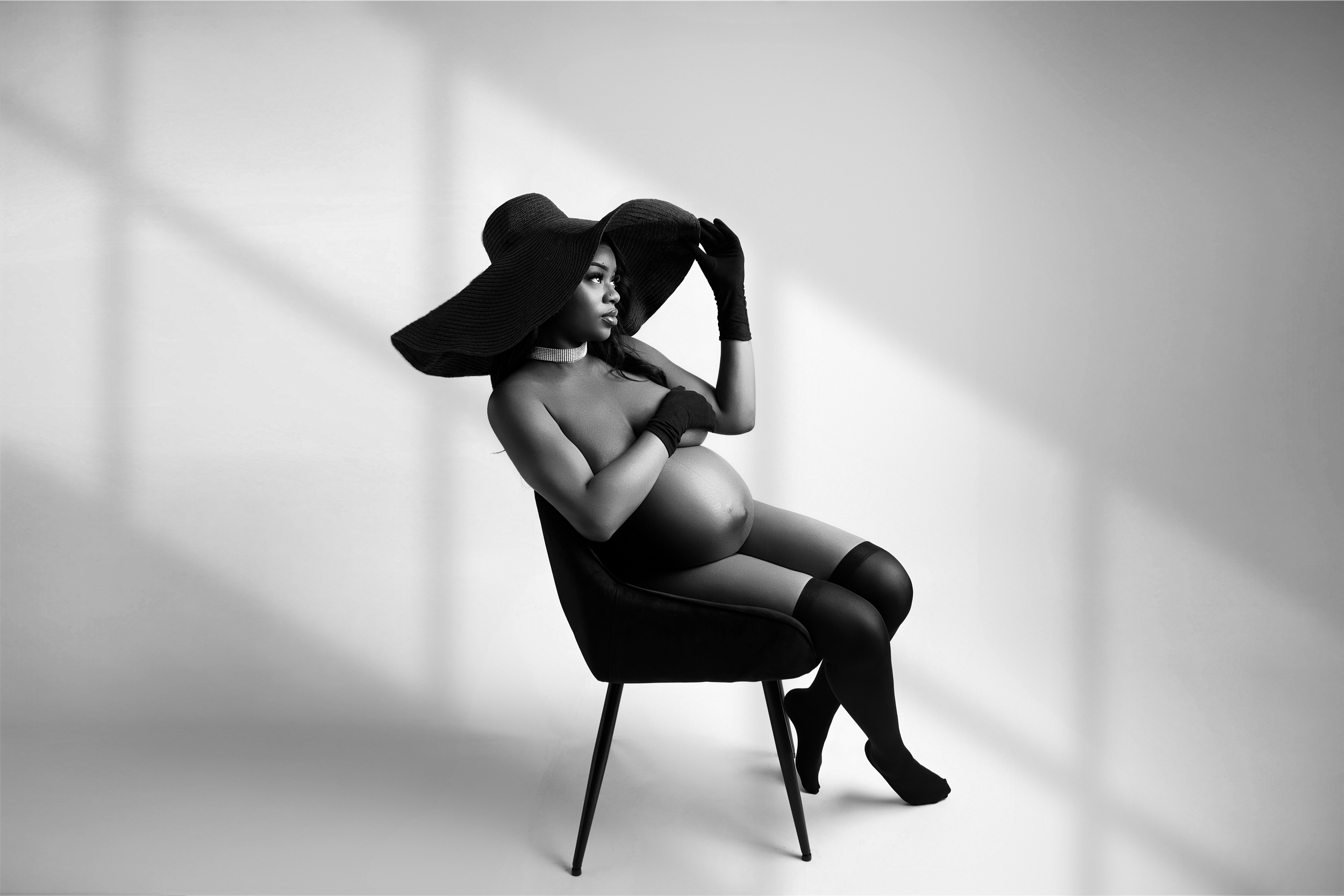 Artistic black and white boudoir photo showing silhouette of person in lingerie and wide-brimmed hat seated on chair.