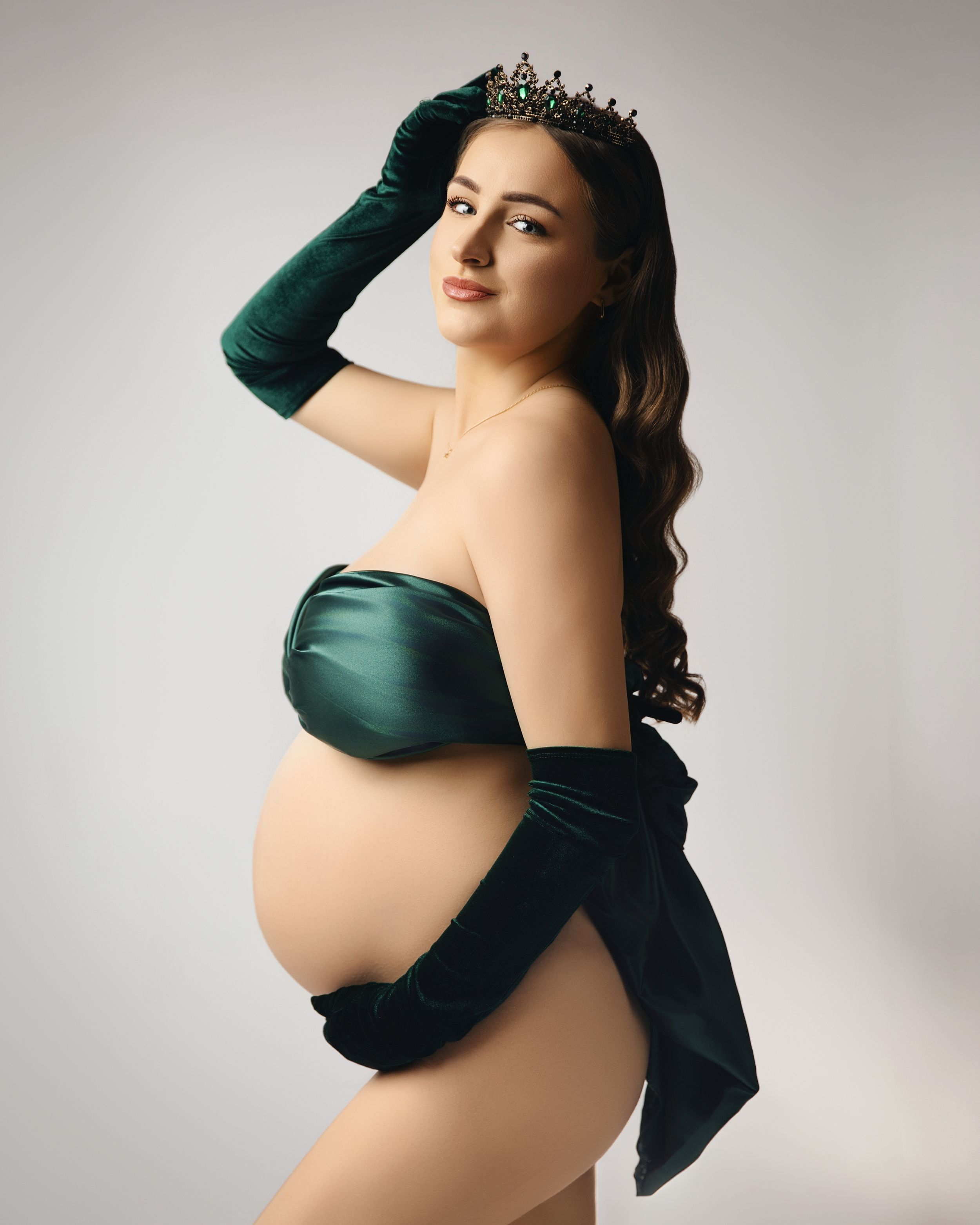 Maternity photo in emerald green gown and tiara showing pregnant belly against light studio background.