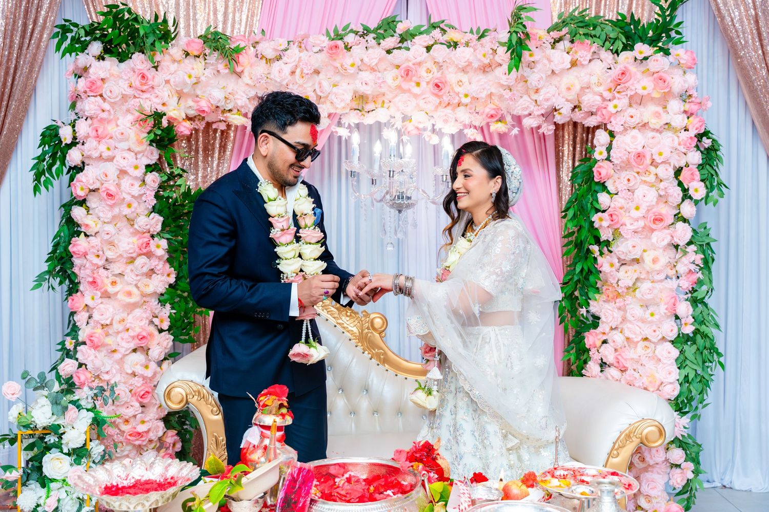 Couple exchanges gifts during wedding celebration under pink floral arch decor.