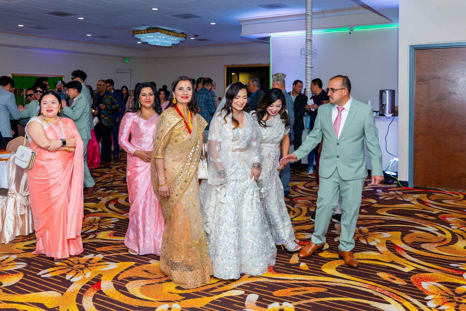 Celebration in a ballroom with guests wearing colorful formal attire on a patterned carpet.
