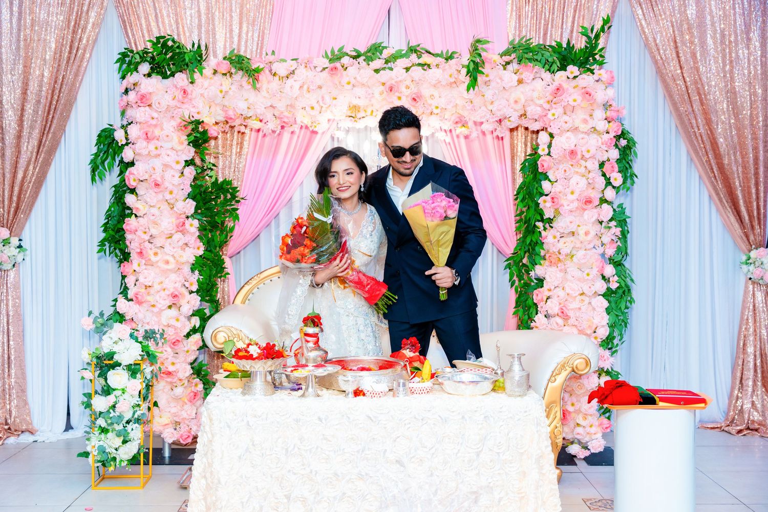 Wedding couple poses at elegant pink floral arch backdrop with party decorations and table setting.