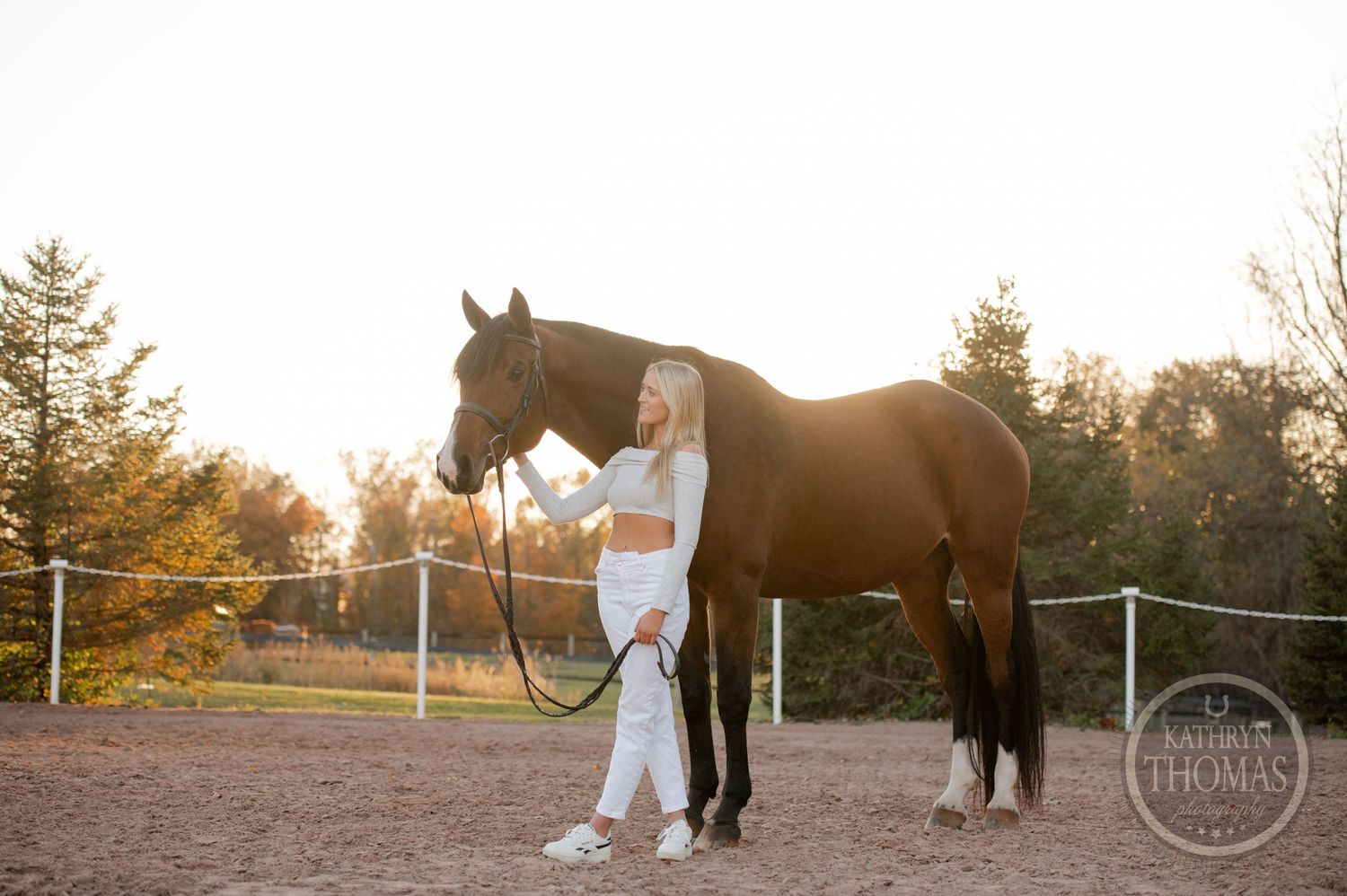 Person in white outfit standing beside brown horse in outdoor arena during golden hour.