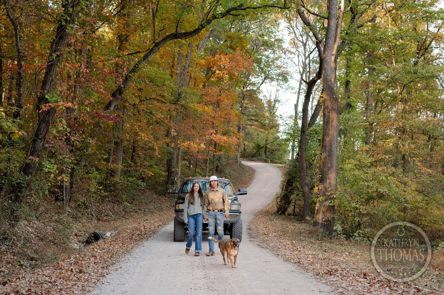 A couple walks their dog down a winding autumn forest path with their truck in the background.
