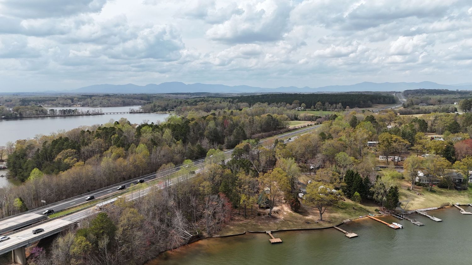 Aerial view of a bridge crossing a lake with residential homes and docks along the wooded shoreline under cloudy skies.