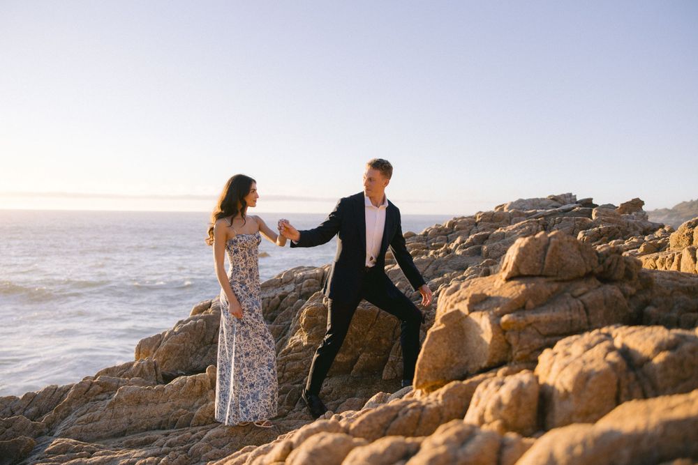 A couple in formal attire holds hands while walking along rocky coastal cliffs at sunset by the ocean.