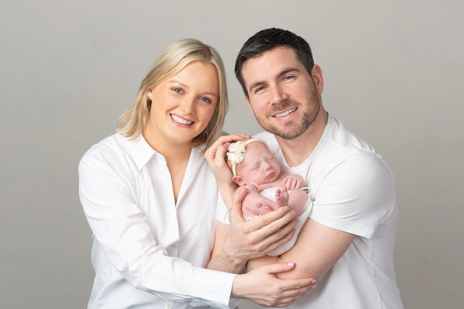 Portrait of new family in white clothing holding newborn baby against grey background.