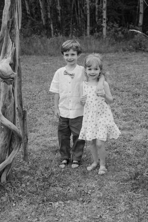 Two small children walk and play together outdoors on a grassy hillside in black and white photography.