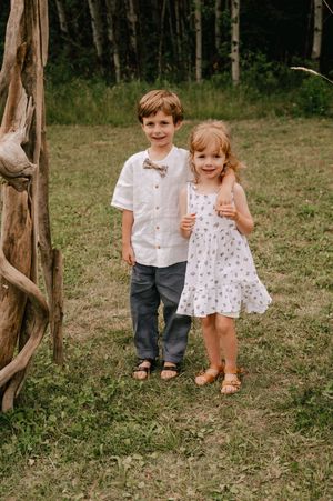 Children in coordinated white summer outfits play together in a wooded outdoor setting.