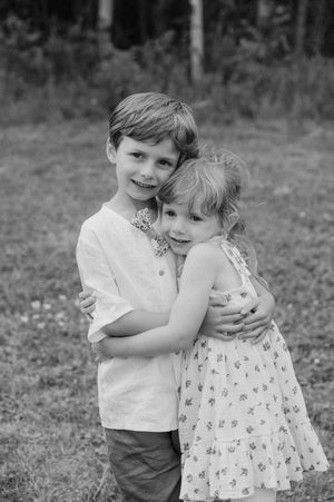 A black and white outdoor portrait shows two young children embracing together in a sweet candid moment.