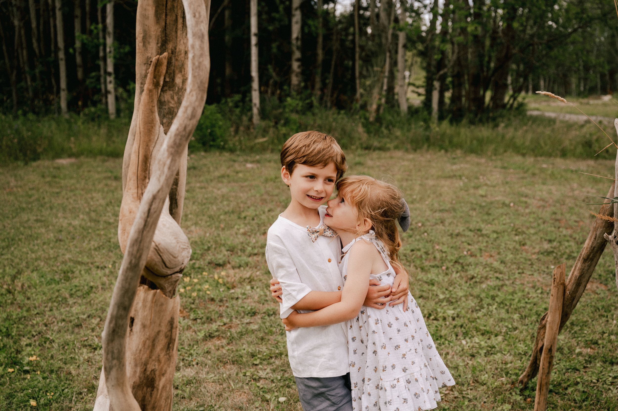 Two children stand together by a tree trunk in a wooded meadow during a summer day.