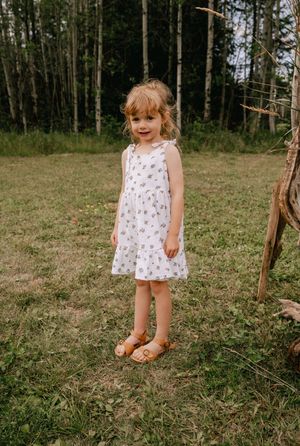 A child in a white floral dress stands playfully in a wooded clearing with sunlight filtering through the trees.