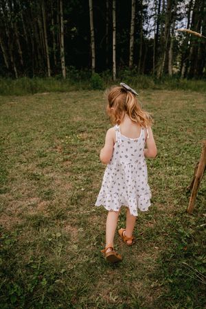 Small child wearing a floral dress and brown shoes walking in a grassy field near tall trees.